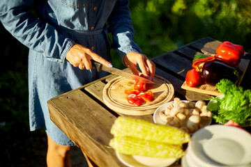 Unrecognizable woman cutting fresh pepper vegetables on wooden board during weekend barbecue in yard, outdoor, prepare for grilling, summer family picnic, food on the nature.