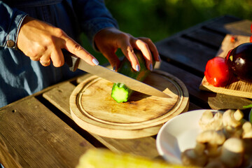 Unrecognizable woman cutting fresh zucchini vegetables on wooden board during weekend barbecue in yard, outdoor, prepare for grilling, summer family picnic, food on the nature.