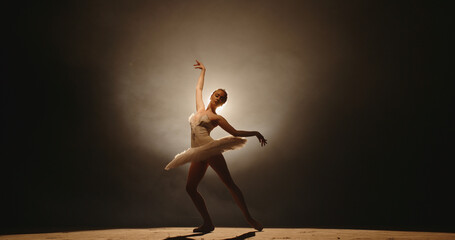 Beautiful caucasian ballerina gracefully dancing and spinning on stage, spotted by white light, isolated on smoked black background 