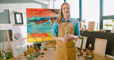 Portrait of joyful smiling young Caucasian professional female artist in apron standing in art studio holding paintbrushes in hands looking at camera in bright workshop just finished work on canvas