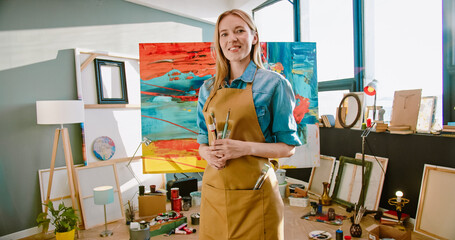 Rear of happy young Caucasian professional female artist in apron standing in art studio looking at...