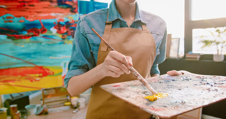 Close up of Caucasian happy positive pretty woman painter using painting tools mixing paints on palette and smiling to camera. Artwork, female artist in workshop, modern art studio, creative concept