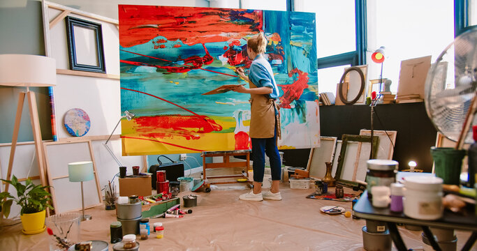 Rear of Caucasian young professional talented female painter working drawing on big canvas in workshop. Modern creating painting on large canva in art studio. Artist in apron working in gallery