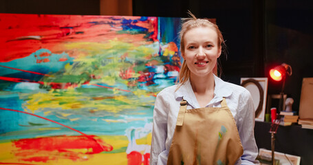 Close up portrait of young Caucasian happy beautiful blonde woman painter looking at camera and smiling after finishing colorful oil painting on large canvas in studio. Art workshop, emotions concept