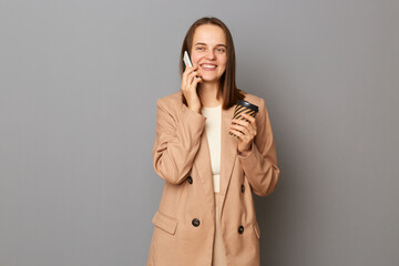 Portrait of cheerful attractive happy woman wearing beige jacket standing isolated over gray background, drinking takeaway coffee and talking on mobile phone, expressing positive emotions.