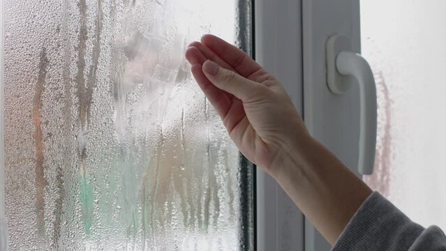 A womans hand touching wet window glass with water drops of condensation. 4K.