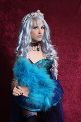 Fototapeta premium Woman With Blue Hair and Blue Corset Dress Holding Blue Fan on Red Velvet Background