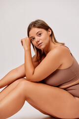 Naklejka premium Vertical shot of body positive confident young woman sits on a floor in studio in beige lingerie, isolated over white background 