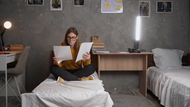 teenager girl student in glasses sits on a bed in a college dormitory holding sheets of paper in her hands reads information. the concept of preparing for the exam.