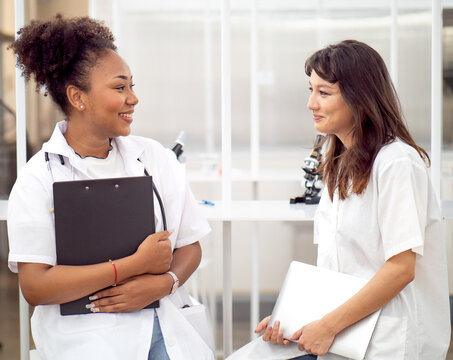 Diverse ethnic medical student women in uniform sitting together talking. Female multiracial doctor student holding document clipboard smiling discussion with a colleague in medical school.