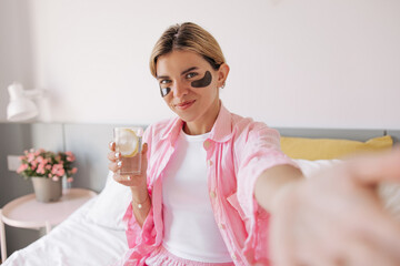 Obraz na płótnie Canvas Beautiful young caucasian woman with eyes patches holds glass of water and looking at camera. Blonde girl wears pajamas, flower in background. Concept perfect start of day.
