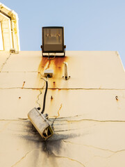 Damaged CCTV camera hanging on a cable on yellow wall, old flood light above the camera. Front element is broken and there is sign of explosion. Blue cloudy sky. War or high criminal unsafe area.