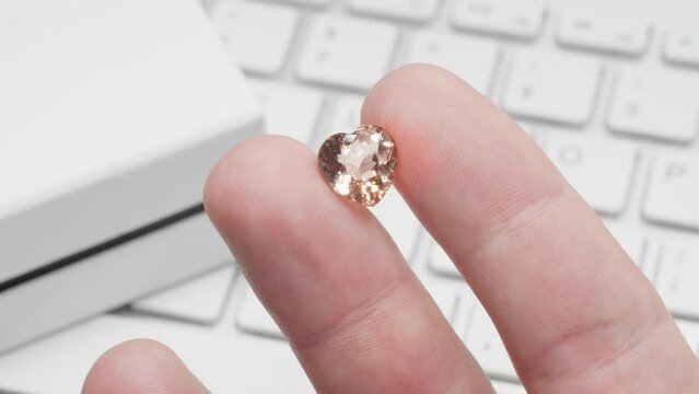 Heart shaped tourmaline gem in hand of gemologist or jeweller. Online jewelry shopping for birthday, Valentine Day, other holiday. Male hand holding peach tourmaline, macro shot with box and keyboard