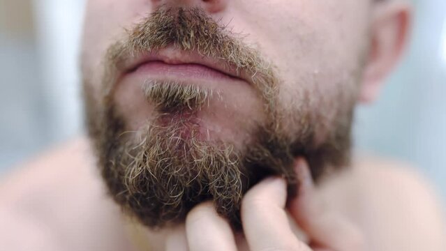 Caring man smoothing his unshaved beard with oil and combing with small brush in the bathroom, daily routine of young handsome man, guy's skincare and grooming concept