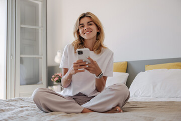Positive young caucasian woman wears casual clothes sitting on bed and using smartphone in morning. Blonde looks at camera with wide smile. Concept of positive emotions, social lifestyle.
