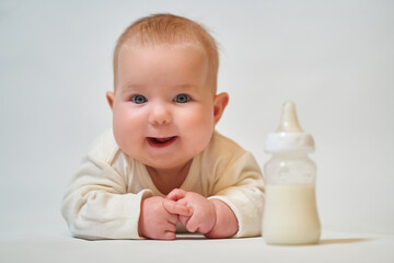 portrait of a smiling baby in light clothes next to a bottle of milk on a light background
