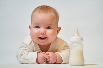 portrait of a smiling baby in light clothes next to a bottle of milk on a light background