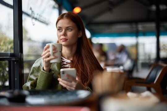 Woman sitting in a cafe drinking coffee from a mug with phone in hand, blogger photographing food content for social media, portrait of a stylish girl with red hair in the autumn