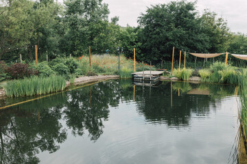 Fototapeta na wymiar Natural swimming pool with biodiversity in countryside resort. Clean pond water with trees reflection and wooden bridge. Rustic getaway for relaxation. Healthy outdoor lifestyle.