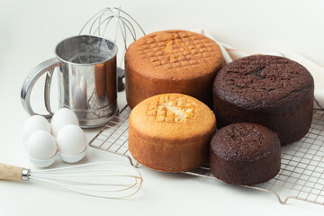 Freshly baked sponge biscuit on a pastry grid next to whisk, sieve for sifting flour and eggs. Brown chocolate and white vanilla sponge cake with professional confectioner utensils. White background