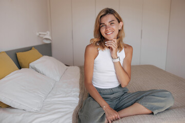 Happy young caucasian woman with blonde hair sitting on bed in bedroom morning. Female dressed casual clothes looking at camera wide smile. Concept positive emotions, weekend home.