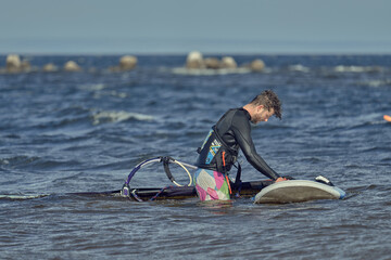 A mature man in a wetsuit, a windsurfer, stands in the water in shallow water near a sailboard.