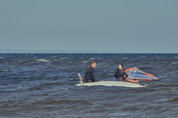 A mature man and a mature woman in wetsuits are standing in the water near the sailboards and talking.