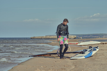 A mature man in a wetsuit, a windsurfer, stands on a sandy shore near a sailboard and rests.