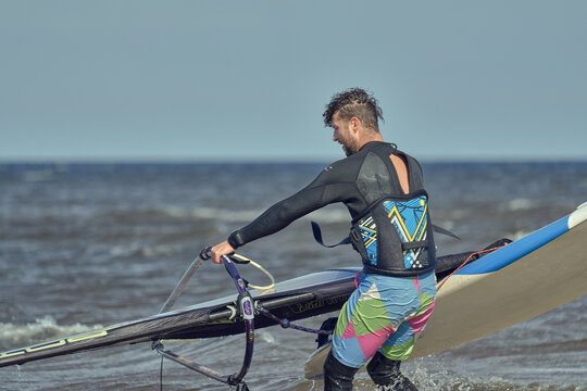A mature man in a wetsuit, a windsurfer, takes a sailboard out of the water on a sunny autumn day. - Powered by Adobe