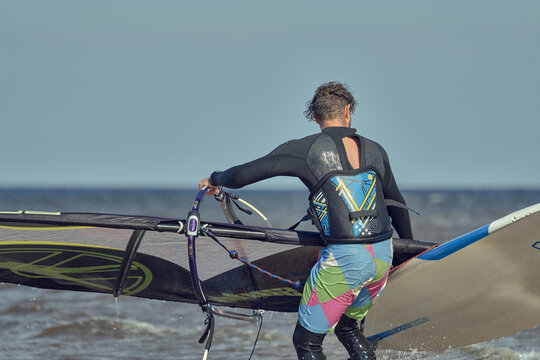 A mature man in a wetsuit, a windsurfer, takes a sailboard out of the water on a sunny autumn day. - Powered by Adobe