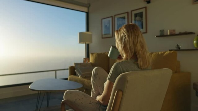 Blonde girl in a cozy sunny apartment drinking morning coffee and watching the sunrise. Woman in her apartment watches the sun rise over the ocean