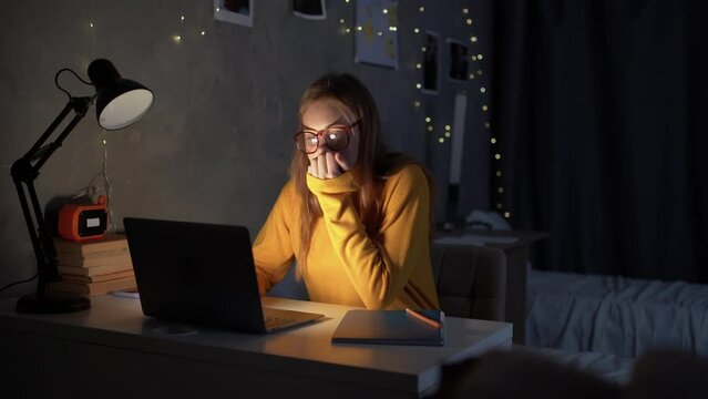 girl teenager student sits at night at a table in a college dormitory looking for information online using a laptop.