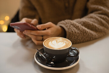 Girl in a fluffy sweater keeps smartphone in hands and writes message next to cup of coffee with...