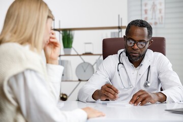 Obraz premium African American doctor, receives a patient, listens to complaints, makes diagnostics. Writes data. Doctor's appointment.