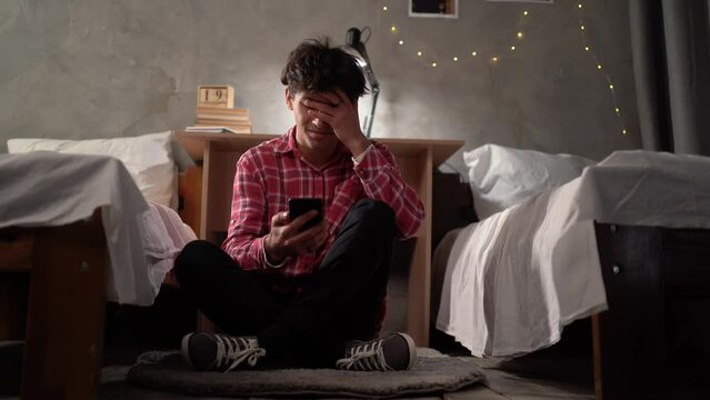 a young student in a college dorm sitting on the floor looking at his smartphone and holding his head, stress and depression. Bullying and blackmail among teenagers.