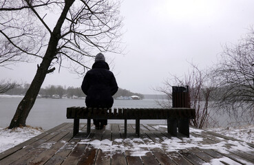 Lonely woman sits on a bench on wood flooring on a winter day and looks at the river view from the...