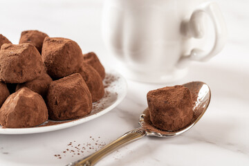 Chocolate truffle on a spoon closeup. Delicious dark chocolate candy balls in cocoa powder and white cup n a marble table. Sweet food, confection and dessert recipe concept. Macro.