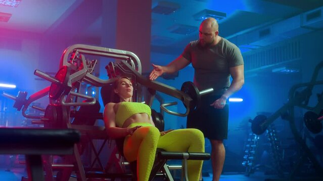 Male trainer teaching woman technique for performing an exercise arm press up on sports simulator. Woman bodybuilder sitting on the machine and doing workout under the guidance of coach in gym.