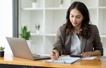 Asian businesswoman sitting happily working on a laptop and taking notes fluently and smiling happily with her assigned tasks at the office.