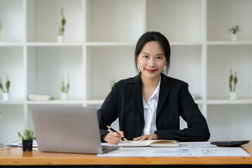 Asian businesswoman sitting happily working on a laptop and taking notes fluently and smiling happily with her assigned tasks at the office.