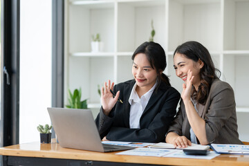 Image of two beautiful Asian businesswomen waving greeting online through a laptop at the table with a calculator, graph, and chart showing business growth data at the office.
