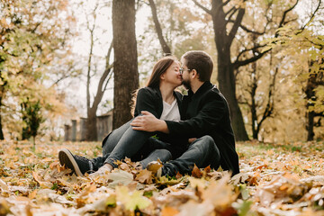 Fototapeta premium Portrait of man kissing with woman in background on sunny autumn park. Tender boyfriend, in casual coat, sitting on ground with beloved woman and kissing during sunny weather