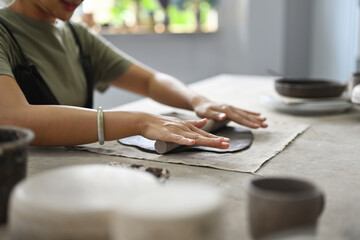 Close up and cropped image with Woman potter master rolling with pin while preparing clay mug,...