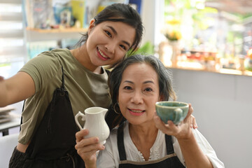 Young and Senior pottery owner take a selfie with product in their workshop studio.