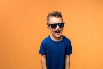 Little boy in sunglasses and blue t-shirt. He smiling, posing on orange background. Childhood,...