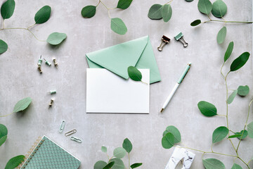 Mint green postal envelope, white card, pen and office stationary. Eucalyptus twigs on stone, overhead view.Delicate green silver dollar eucalyptus leaves. Trendy, modern birthday greeting mockup.