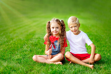 Naklejka na ściany i meble children girl and boy blonde with a big lollipop heart in the summer on the lawn on the green grass, the concept of the valentine's day holiday, space for text