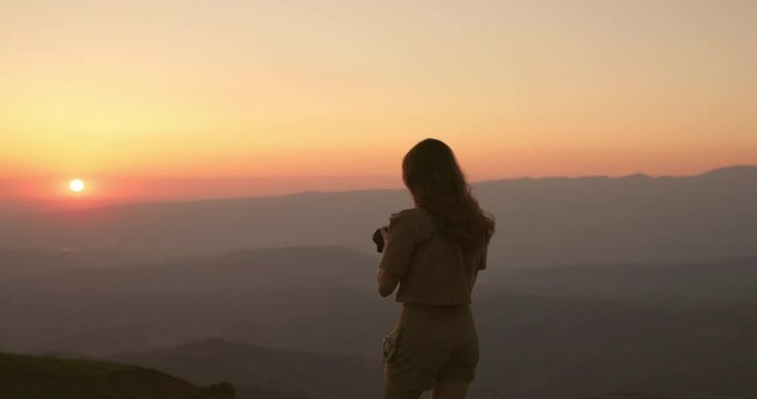 Shot from behind, young female traveler with long hair takes photographs of a hilly valley with a professional camera. Woman takes pictures at sunset. Travel and hiking in the mountains for the photos