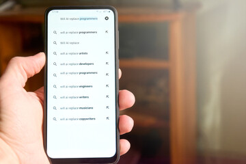 Male hand holding mobile phone screen with search "Will AI replace programmers" and list of suggested searches below. People searches on AI, concept of fear about AI replacing jobs. Social issue.