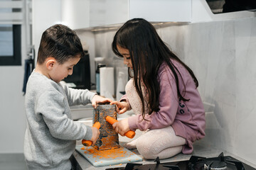 Brother and sister cook in the kitchen together, grating carrots. Children help mother, cooking healthy food at home. Lived-in Interior, real life. Healthy organic food.